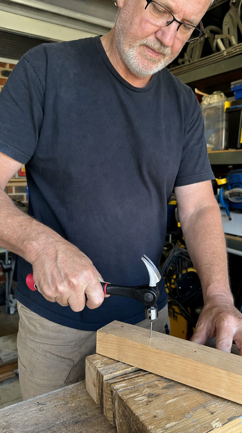 A man with glasses starting a nail in a timber block with the claw hammer